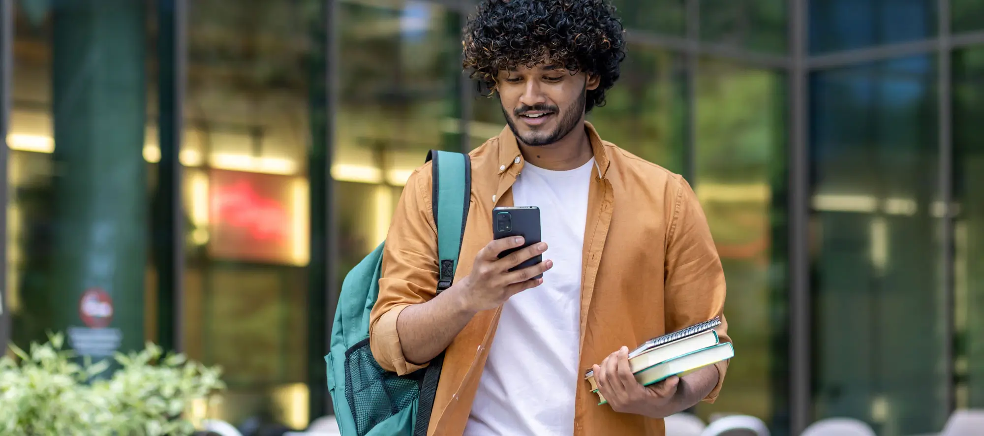 Young male student walking on street near campus with backpack. Holds books and uses a mobile phone.