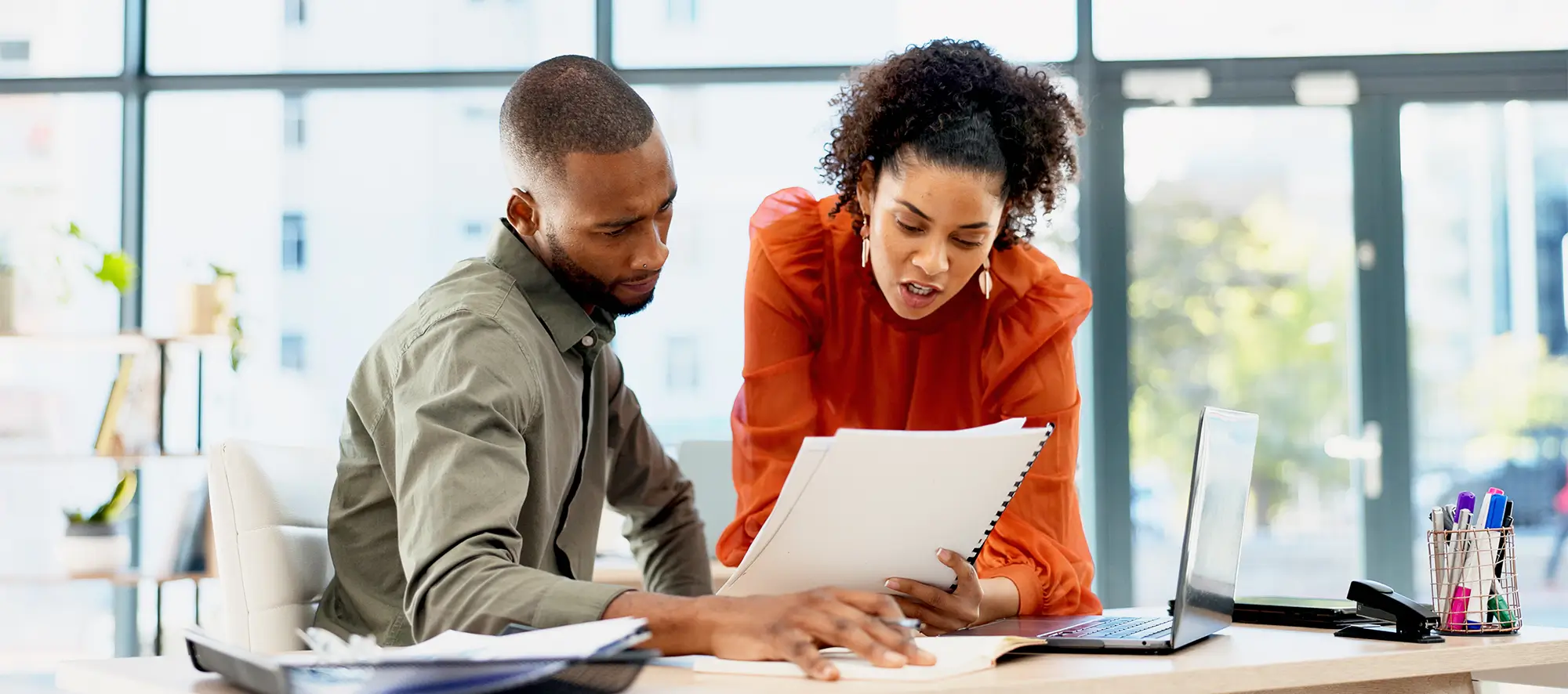 Two employees talking about paperwork at a desk.
