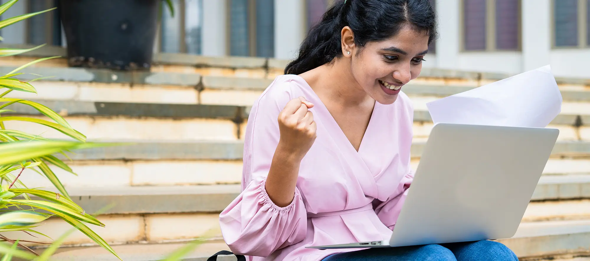 Student expressing excitement while looking at a laptop on campus stairs.