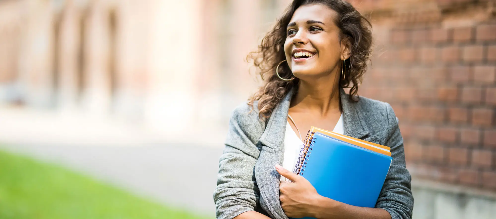 Pretty cheerful latin american student smiling at camera carrying notebook on campus at college