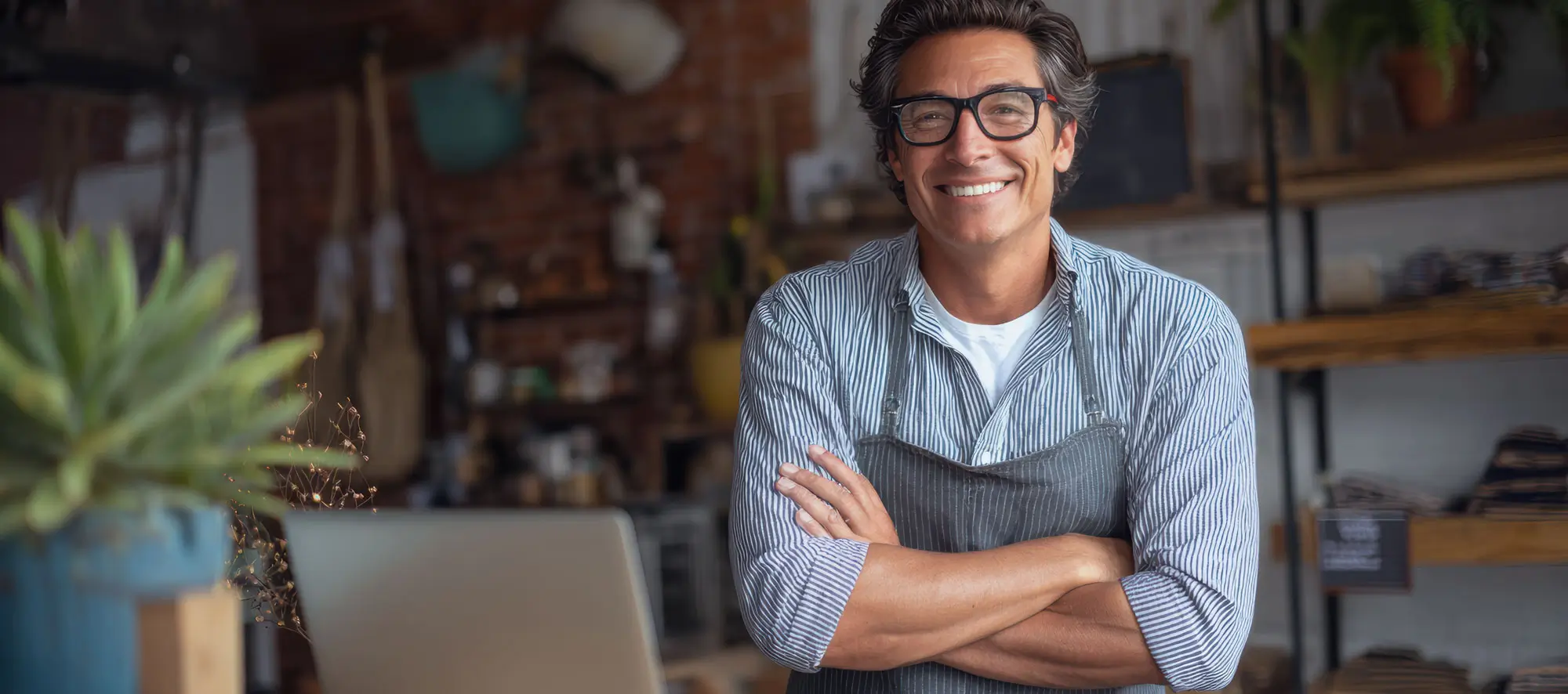 Confident hispanic male adult entrepreneur in apron with laptop in cozy store