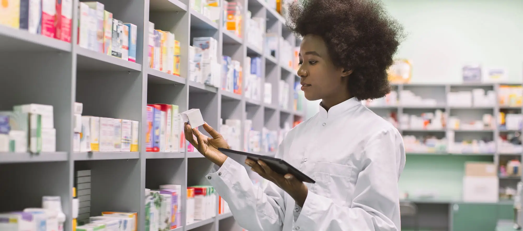 Female pharmacist using digital tablet, while standing near the shelves with medicines in modern pharmacy during drugs inventory.