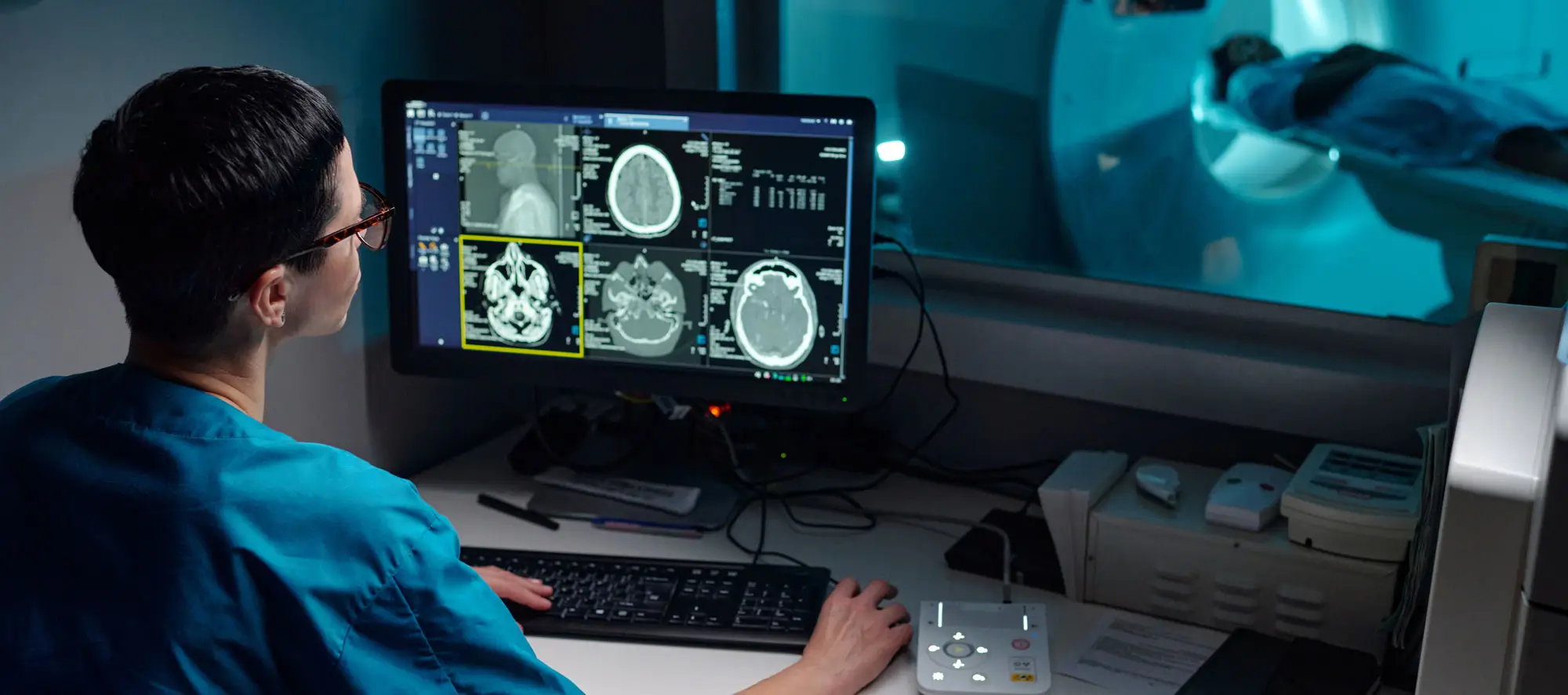 Medical professional reviewing patient's MRI images on computer monitor with patient in MRI machine in background.