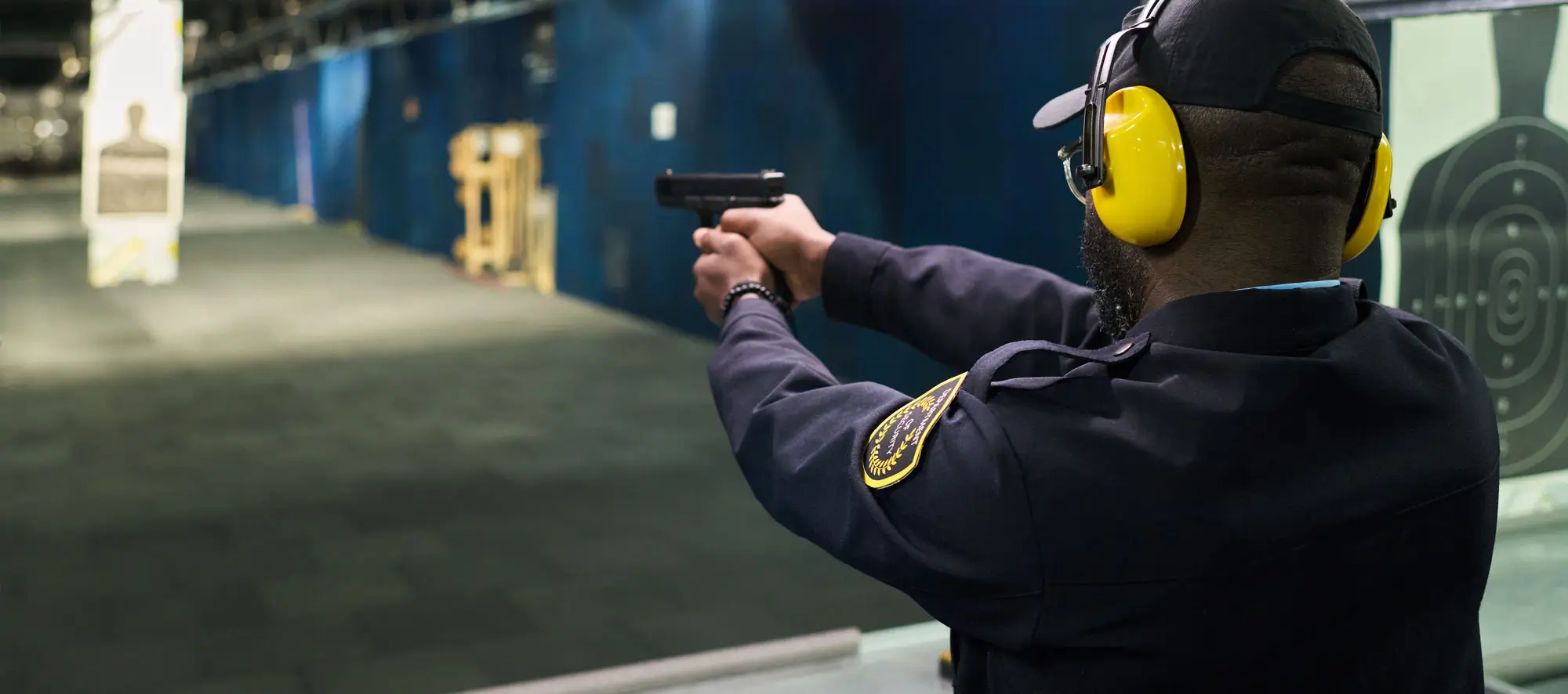 Person wearing safety gear practicing shooting at indoor gun range aiming at target downrange. Protective earmuffs and glasses used for hearing and eye protection