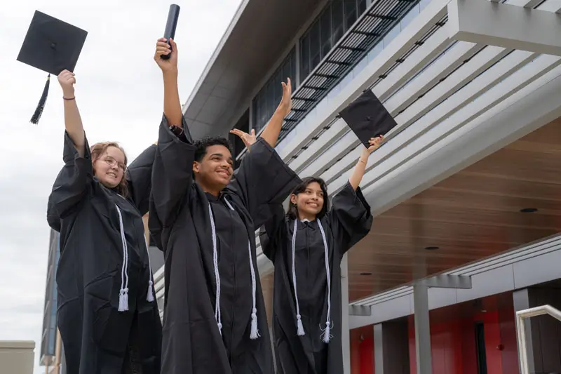 Three students in regalia with arms raised in the air, smiling.