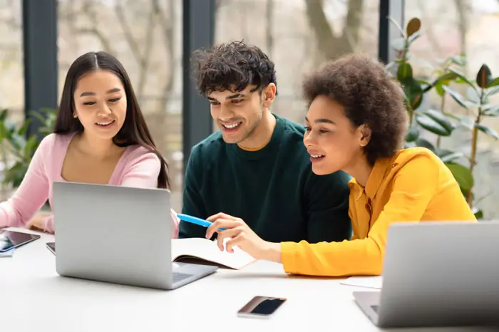 Happy international students friends sitting at desk in coworking space and using laptop