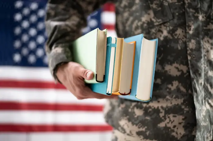 Military person holding a stack of books in front of the American flag.