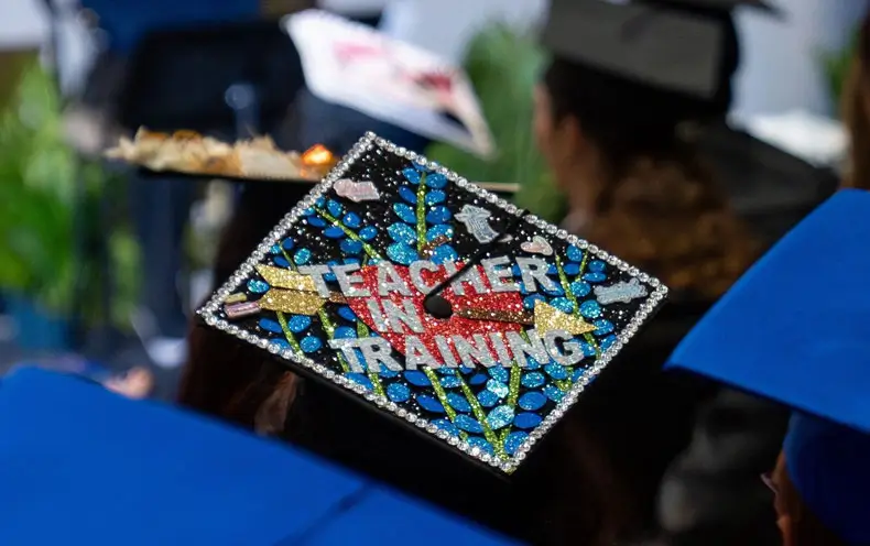 A COM Graduate's decorated cap reads 'Teacher in training'