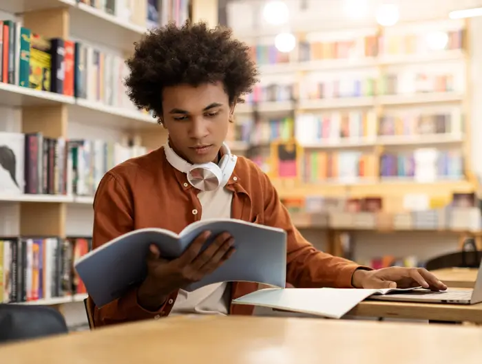 Student in library reading a textbook while working on a laptop.