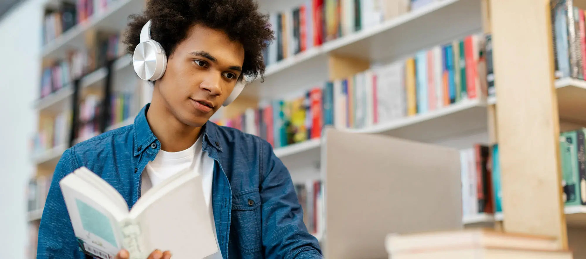 Focused black male college student wearing headphone, sitting at desk in library, studying, holding book and using laptop for research or project